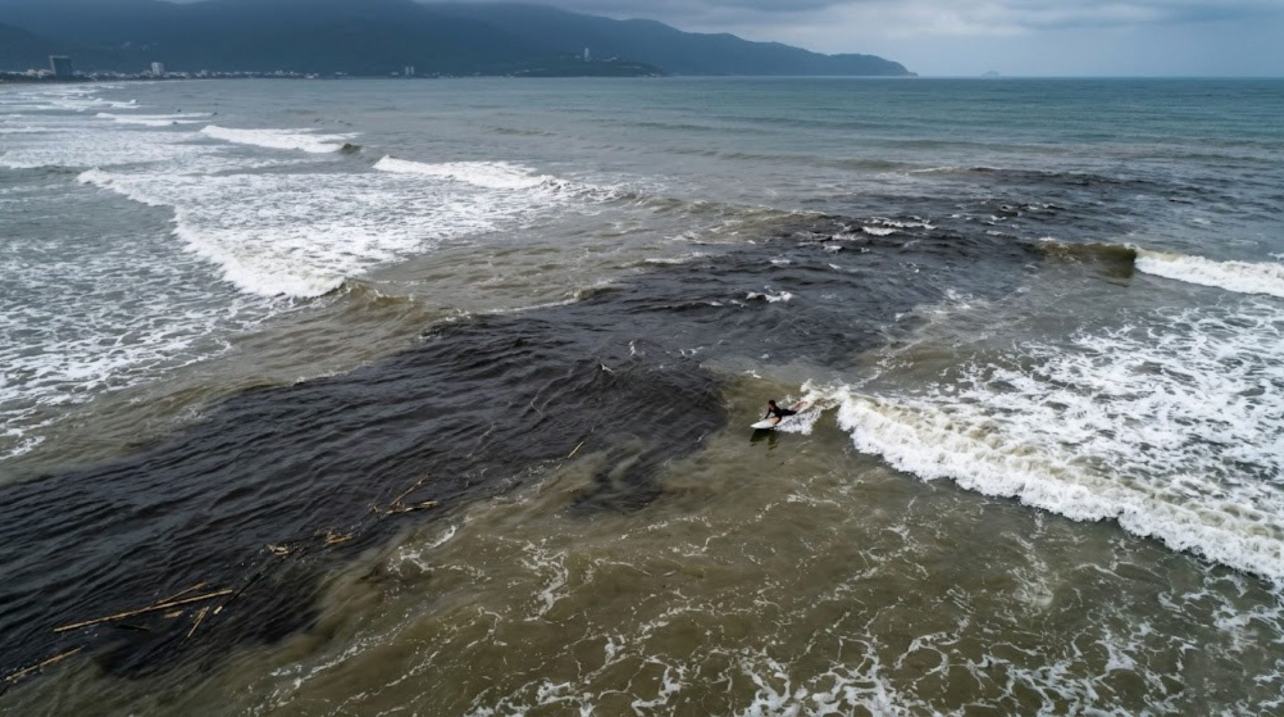 An aerial drone photograph taken over My Khe Beach, Da Nang, showing a powerful rip current channel. A distinctly darker, flat river-like channel of water flows straight out to sea between whitewater breaking zones. Dark murky water and bamboo debris are visible in the foreground. A lone surfer on a board paddles within the dark water, illustrating a dangerous paradoxical zone. The moody Son Tra Peninsula is in the background