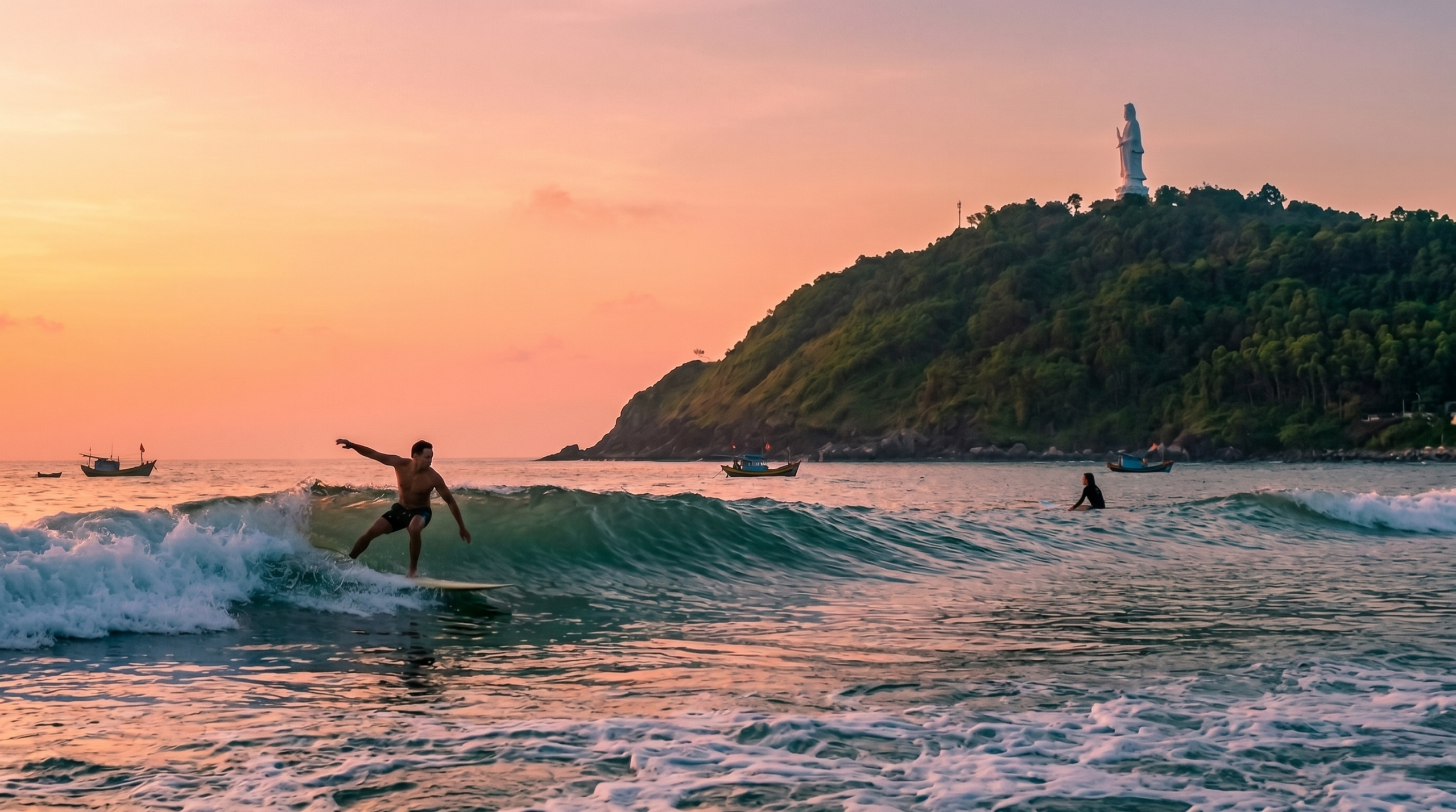 Sunset surfing on My Khe waves in Da Nang. In the left foreground, a man balances on his surfboard riding a wave. In the midground, a woman paddles on her board. The background features the lush green Son Tra Peninsula crowned by the giant white Lady Buddha statue, with a few fishing boats anchored under an orange-pink sky
