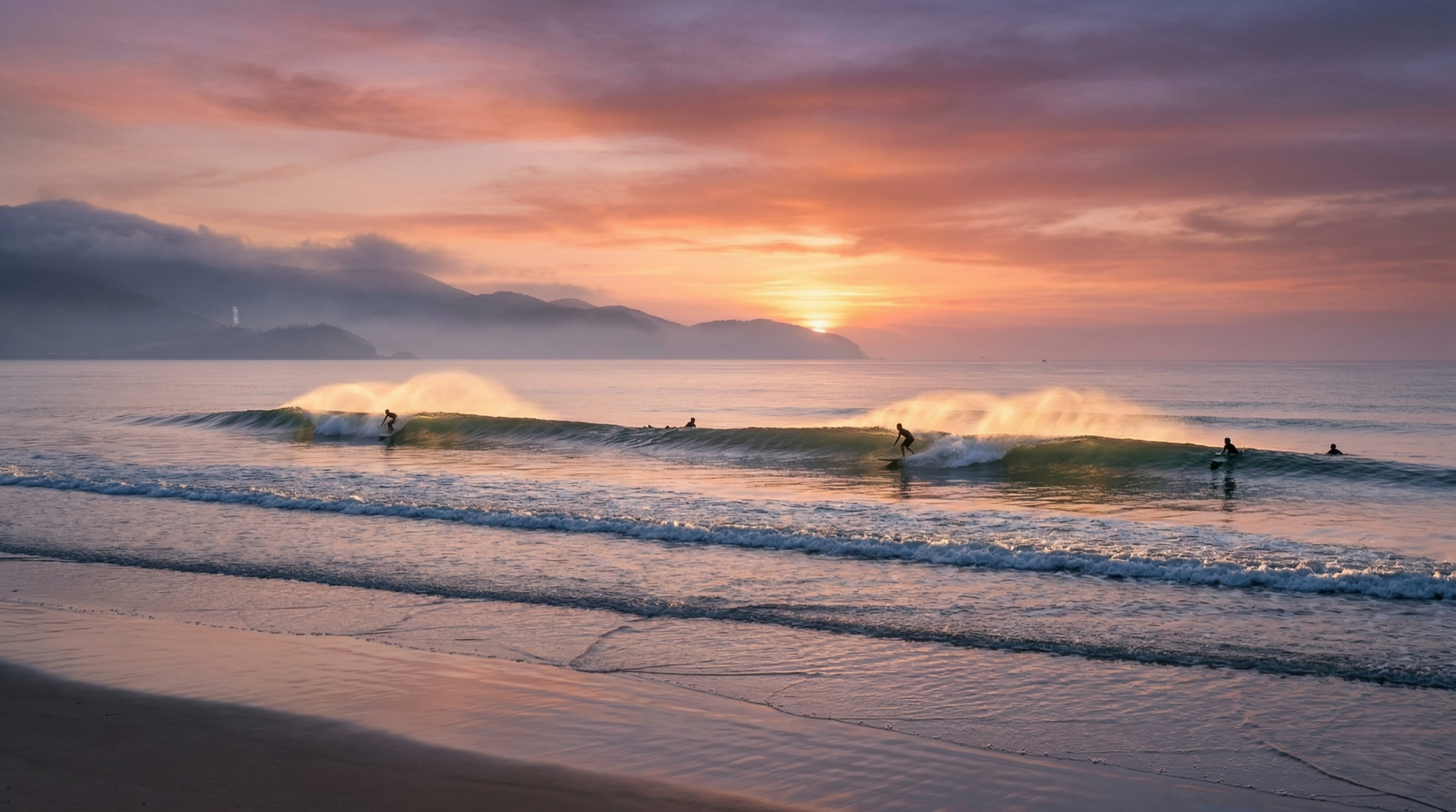 Wide shot of surfers riding long, smooth waves at My Khe beach, Da Nang under a vibrant pink and orange sunrise sky, with the misty Son Tra peninsula in the background.